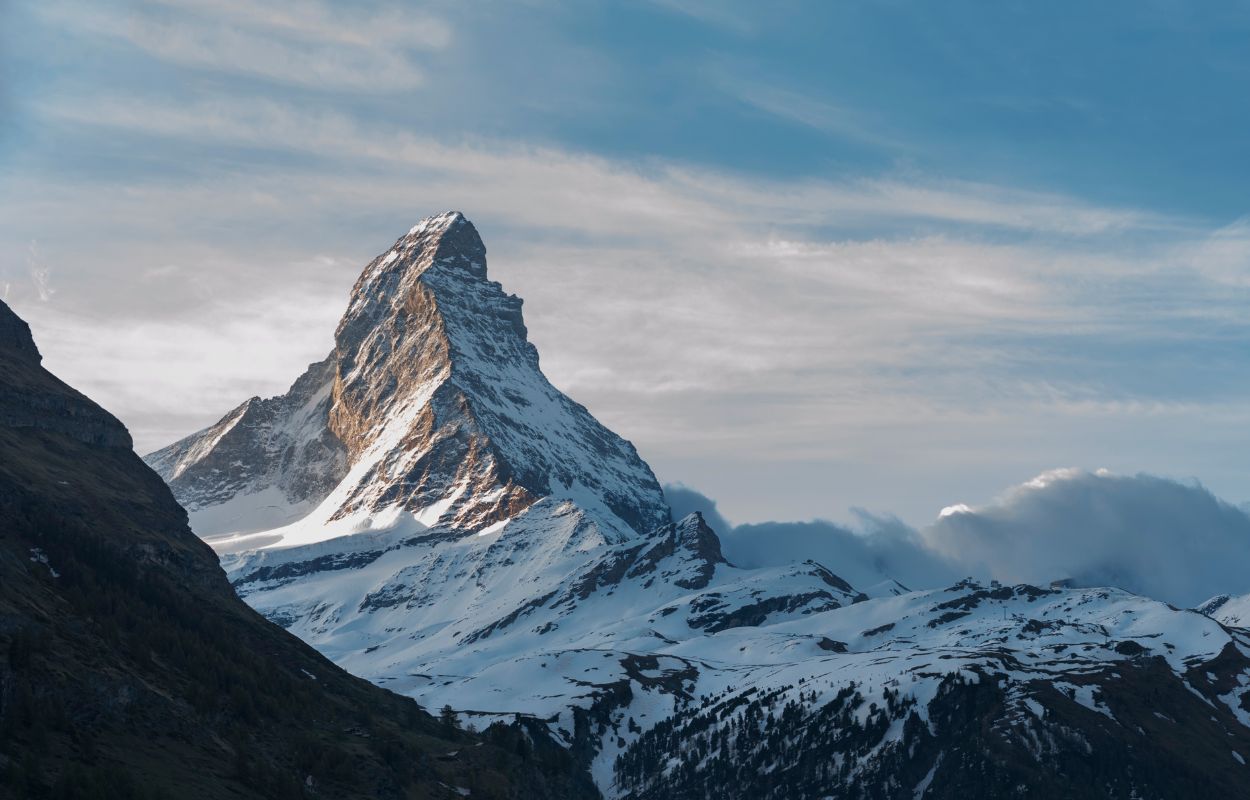 Snow-covered Matterhorn mountain peak in the Swiss Alps, lit by soft sunlight with dramatic clouds and a blue sky in the background. Lower slopes show patches of forest and rocky terrain