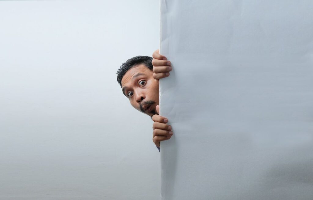 A surprised man peeking from behind a white wall or panel, with wide eyes and raised eyebrows, creating a humorous or curious expression.