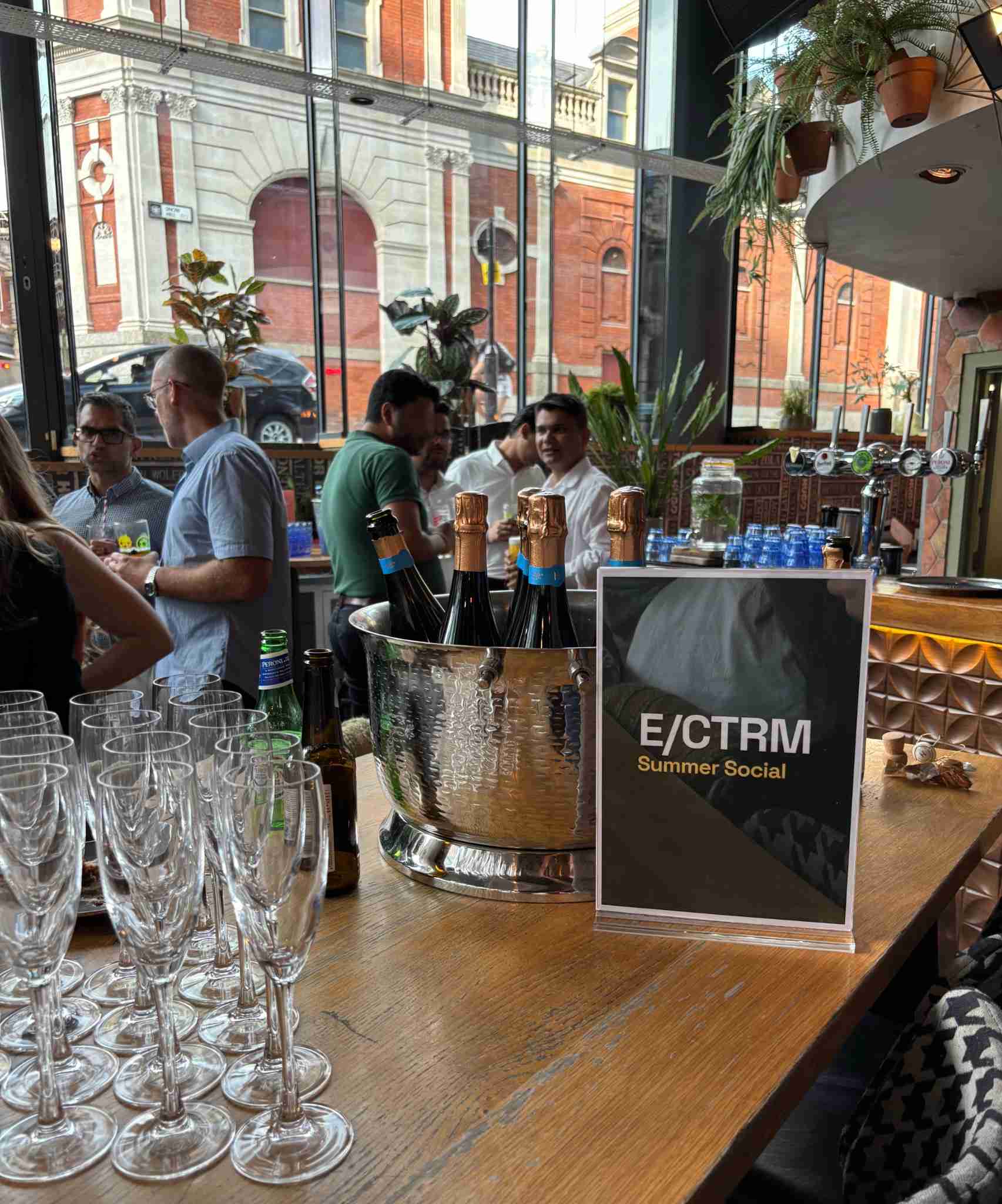 People gathered at a summer social event in a modern bar with large windows, bottles of champagne in a silver ice bucket, and a sign reading 'E/CTRM Summer Social' on the wooden bar counter alongside rows of empty champagne glasses.